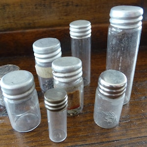 May include: Seven small glass bottles with silver lids. The bottles are clear and contain various items, such as beads and small objects.