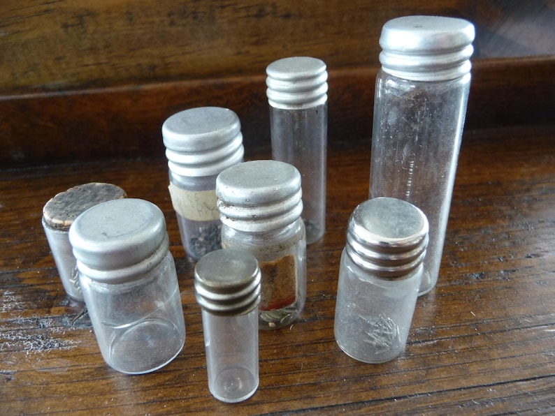 May include: Seven small glass bottles with silver lids. The bottles are clear and contain various items, such as beads and small objects.