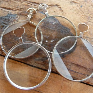 May include: Four silver-toned magnifying glasses with round lenses and a hinge on each side. The magnifying glasses are on a wooden surface.
