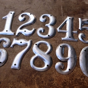 May include: Ten silver metal house numbers, 1 through 10, with a vintage style. Each number has a slightly different design and is slightly worn.
