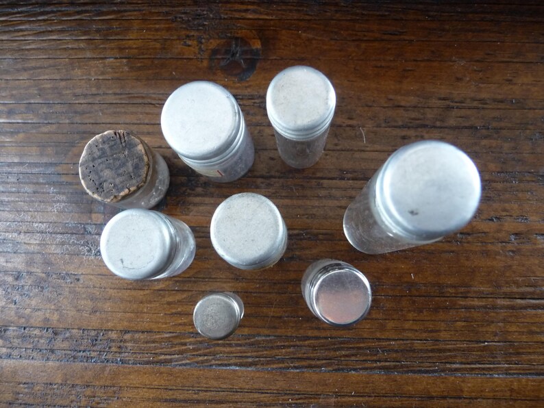 May include: A collection of eight small, clear glass bottles with silver lids. The bottles are arranged in a circle on a wooden surface.
