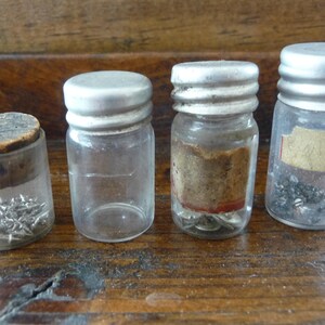 May include: Four small glass jars with silver lids, each containing different small metal objects. The jars are on a wooden surface.