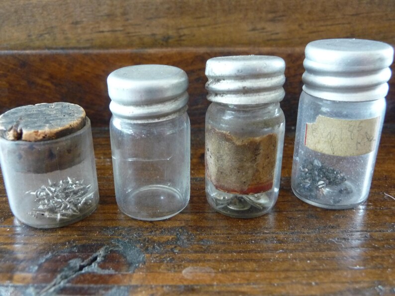 May include: Four small glass jars with silver lids, each containing different small metal objects. The jars are on a wooden surface.