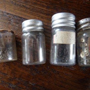 May include: Four small glass jars with metal lids, filled with various small objects. The jars are arranged in a row on a wooden surface.