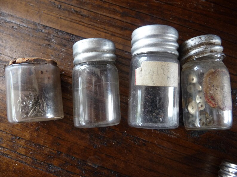 May include: Four small glass jars with metal lids, filled with various small objects. The jars are arranged in a row on a wooden surface.