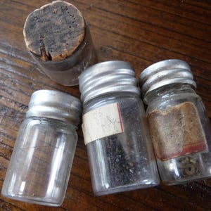 May include: Four small glass jars with silver lids. The jars are filled with various items, including small black seeds, metal pieces, and a cork stopper.
