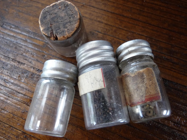 May include: Four small glass jars with silver lids. The jars are filled with various items, including small black seeds, metal pieces, and a cork stopper.