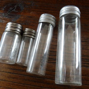 May include: Four clear glass bottles with silver lids. The bottles are different sizes and are arranged in a row from largest to smallest.