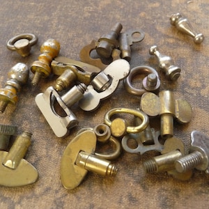 May include: A collection of antique metal hardware pieces, including knobs, screws, and keys, in various shapes and sizes. The hardware has a tarnished, aged appearance with brass, silver, and bronze tones. The items are scattered on a textured, brown surface.