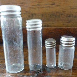 May include: Four clear glass bottles with silver lids of varying sizes. The bottles are empty and have a slightly frosted appearance.