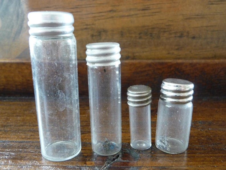 May include: Four clear glass bottles with silver lids of varying sizes. The bottles are empty and have a slightly frosted appearance.
