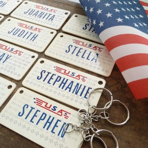May include: Cream-colored keychains with names like "Ray" and "Stephen" are displayed, each featuring a USA logo and state abbreviations. An American flag is in the background, with a few keychains attached to a metal ring.