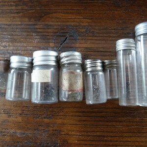 May include: Eight small glass jars with silver lids, some containing small metal objects. The jars are arranged in a row on a wooden surface.