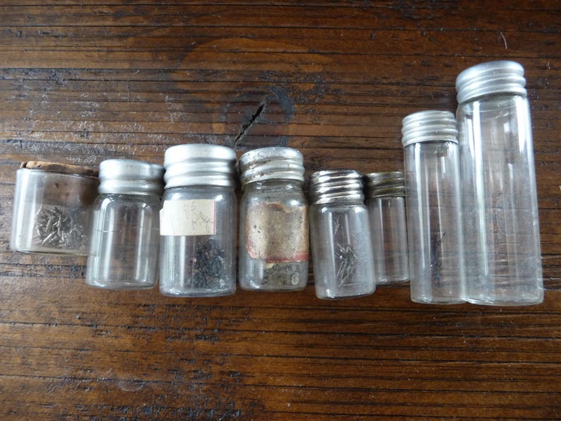 May include: Eight small glass jars with silver lids, some containing small metal objects. The jars are arranged in a row on a wooden surface.