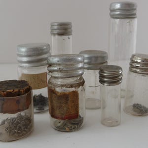 May include: A collection of eight small glass jars with silver lids. The jars are filled with various small objects, such as beads, buttons, and other trinkets.