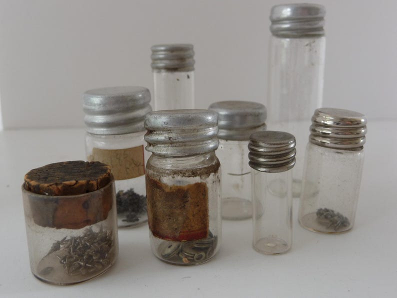 May include: A collection of eight small glass jars with silver lids. The jars are filled with various small objects, such as beads, buttons, and other trinkets.