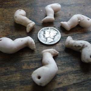 May include: Six antique porcelain doll legs in a circle around a silver coin. The legs are off-white with some discoloration and small holes. The coin has a profile of a person on it.