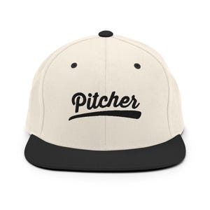 Könnte beinhalten: Cremefarbene Baseballkappe mit schwarzem Schirm. Das Wort "Pitcher" ist in schwarzer Schrift auf der Vorderseite gestickt. Die Kappe hat einen flachen Schirm und einen Knopf oben.