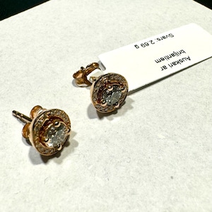 Puede incluir: Un par de pendientes de oro con un diamante redondo central rodeado de diamantes más pequeños. Los pendientes están sobre un fondo blanco con una etiqueta que dice "Auskarat Diamanten  Svars: 2,69 g".