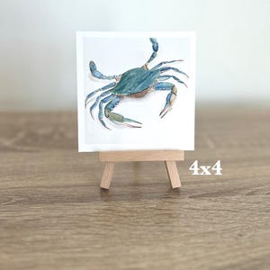 May include: A watercolour painting of a blue crab on a white background, displayed on a small wooden easel. The artwork is 10x10 cm. The crab has detailed claws and legs, with a light brown wooden easel.