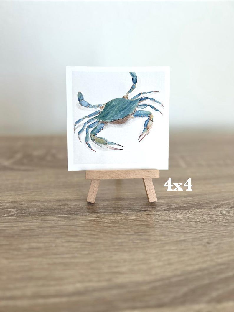 May include: A watercolour painting of a blue crab on a white background, displayed on a small wooden easel. The artwork is 10x10 cm. The crab has detailed claws and legs, with a light brown wooden easel.