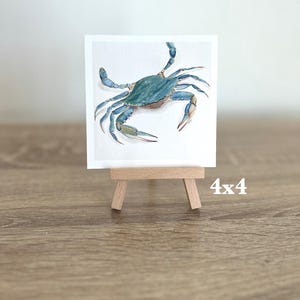 May include: A watercolour painting of a blue crab on a white background, displayed on a small wooden easel. The artwork is 10x10 cm. The crab has detailed claws and legs, with a teal and blue colour palette.