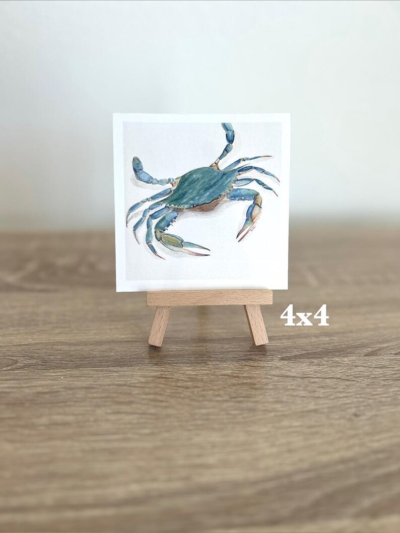 May include: A watercolour painting of a blue crab on a white background, displayed on a small wooden easel. The artwork is 10x10 cm. The crab has detailed claws and legs, with a teal and blue colour palette.