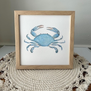 May include: A square framed watercolor painting of a blue crab centered on a white background. The crab features shades of light and medium blue with detailed pincers and legs. The artwork is enclosed in a light-toned wooden frame with a simple profile.