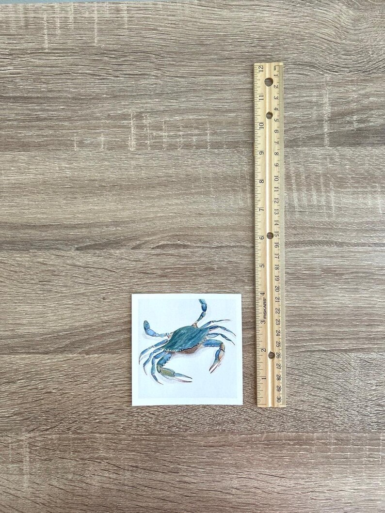May include: A square ceramic tile with a blue crab illustration. The crab is rendered in shades of blue and green. A wooden ruler, marked in inches, is next to the tile. The tile is approximately 7.6 cm by 7.6 cm.