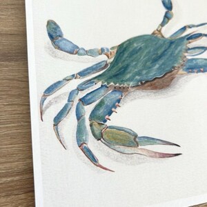 May include: Watercolour painting of a blue crab. The crab has a teal-coloured shell and blue legs and claws. The artwork is on white paper, with a wooden surface visible in the corner.