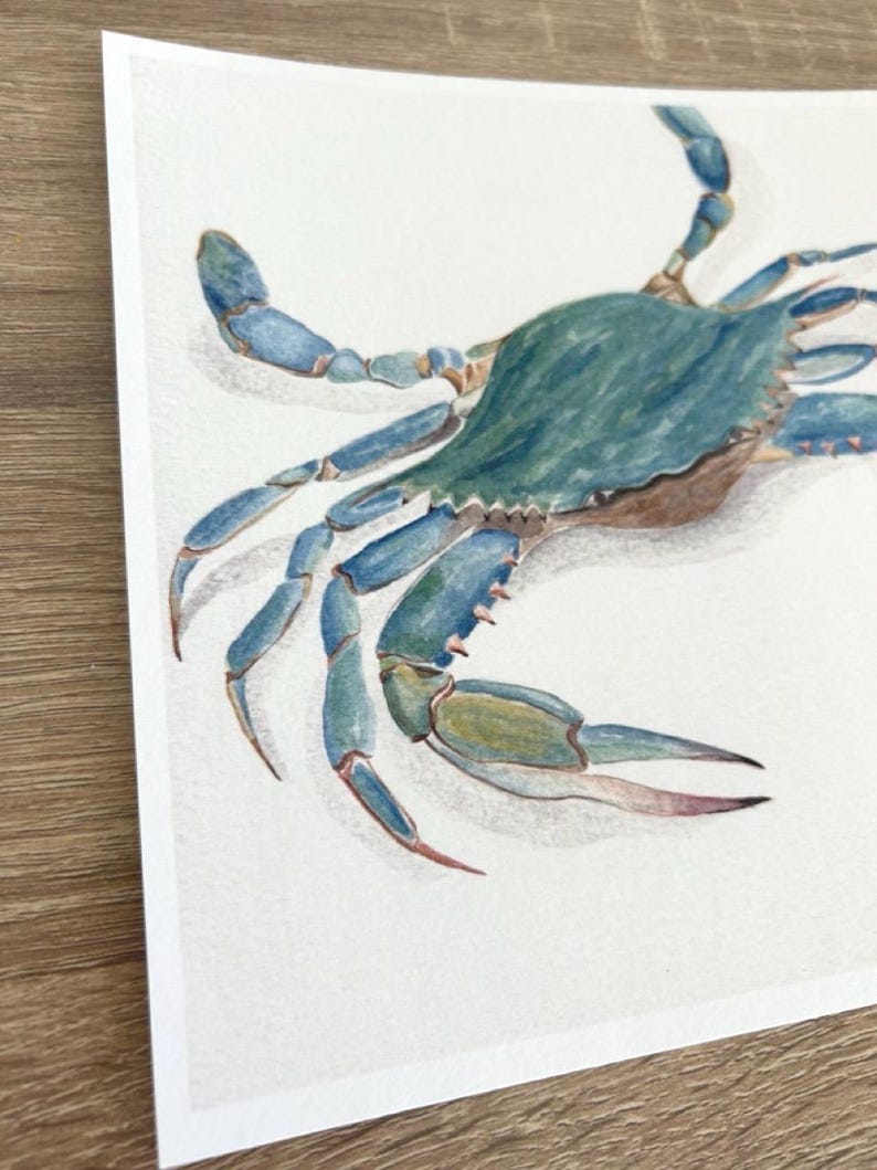 May include: Watercolour painting of a blue crab. The crab has a teal-coloured shell and blue legs and claws. The artwork is on white paper, with a wooden surface visible in the corner.