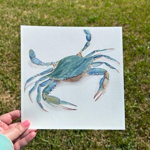 May include: A watercolour painting of a blue crab. The crab has a blue-green carapace and blue legs and claws. The artwork is on a white square paper, held by a person with pink nail polish, against a green grass background.