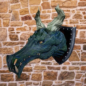 May include: A detailed dragon head wall sculpture. The dragon is primarily dark green and blue with yellow eyes and light green horns. It is mounted on a black shield with silver rivets, set against a stone wall.