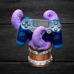 May include: A blue and black video game controller is held by purple tentacles emerging from a wooden barrel. The controller has blue accents and buttons. The barrel is brown with silver studs.