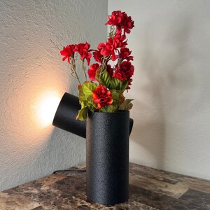 May include: A black, textured, cylindrical lamp with a spotlight feature, holding vibrant red flowers and green leaves. The lamp is on a marble-patterned surface, casting a warm glow.