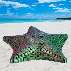 May include: A large, textured, iridescent green and purple starfish rests on a sandy beach. The starfish has a scale-like pattern and is set against a backdrop of a blue sky, white sand, and ocean.