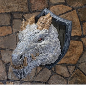Peut inclure: Sculpture détaillée d'une tête de dragon avec une finition texturée grise et blanche, montée sur un bouclier. Le dragon présente des cornes et des défenses dorées, et un œil rouge et jaune. Le bouclier a un cadre en métal foncé.
