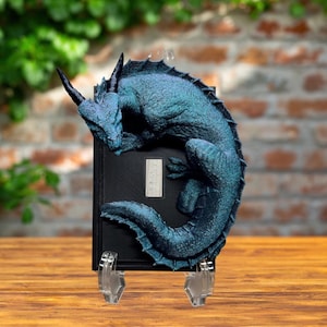 May include: A teal and blue dragon sculpture perched on a black book. The dragon has horns and a textured body. The book is held up by a clear stand, and the background is a brick wall and greenery.