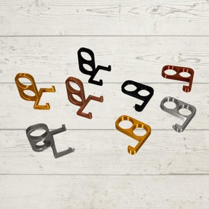 May include: A collection of multi-colored, metal, no-touch door openers. The openers are in various colors, including gold, black, bronze, and silver. Each opener has a finger hole and a hook for opening doors and pressing buttons.