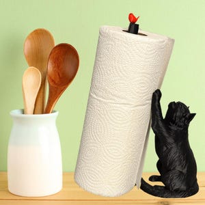 May include: A black cat-shaped paper towel holder with a roll of white paper towels. A small red bird sits atop the holder. A white ceramic jar holds wooden spoons of varying sizes.