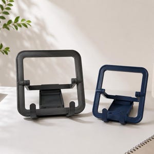 May include: Two plastic phone stands, one dark gray and one navy blue, are displayed on a white surface. Each stand has a square frame and an adjustable base with hooks. Pens and greenery are in the background.