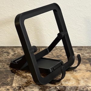 Puede incluir: Un soporte plegable negro para tabletas. El soporte está hecho de un material negro mate y presenta una base rectangular con dos brazos curvos para sujetar la tableta. El soporte tiene un marco cuadrado y un diseño articulado.