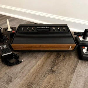 May include: An Atari video computer system console featuring a black ribbed top and a wood-grain front panel with the Atari logo. The set includes a black power adapter and four black joysticks, two of which are labeled Starfighter.