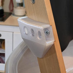 以下が含まれることがあります： 3つの楕円形の開口部を持つ半透明の3Dプリント製プラスチックブラケット。金属製のネジでハイチェアの明るい色の木製フレームにしっかりと固定されています。ブラケットは先細りの角張ったデザインで、表面は質感のある仕上げです。