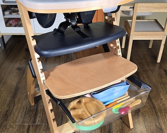 May include: A wooden high chair with a black seat and tray. The chair has a clear plastic tray and a storage bin with bowls, spoons, and a blue container. The chair is on a wood floor.