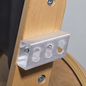 Op de afbeelding: Close-up van een houten stoelframe met een metalen beugel bevestigd. De beugel is zilverkleurig en heeft drie gaten. Het hout is lichtbruin en de schroeven zijn zilver.