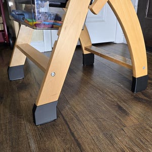 May include: A wooden high chair with a clear plastic tray and storage container. The chair has a natural wood finish with black accents on the feet. The chair is on a dark brown wood floor.