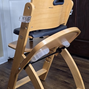 Può includere: Un seggiolone in legno con schienale curvo e vassoio. La sedia ha una finitura in legno naturale con dettagli neri e grigi. La seduta e lo schienale sono imbottiti. La sedia è progettata per un bambino.