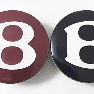 May include: Two round, glossy buttons. One is a deep burgundy color, the other is black. Each button features a large, white letter "B" in a bold, serif font. The buttons are set against a white background.