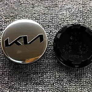 Puede incluir: Un tapacubos plateado brillante de Kia con el logotipo de Kia en negro. Junto a él, una tapa de plástico negro con el texto "XW0609-S" y otras marcas.